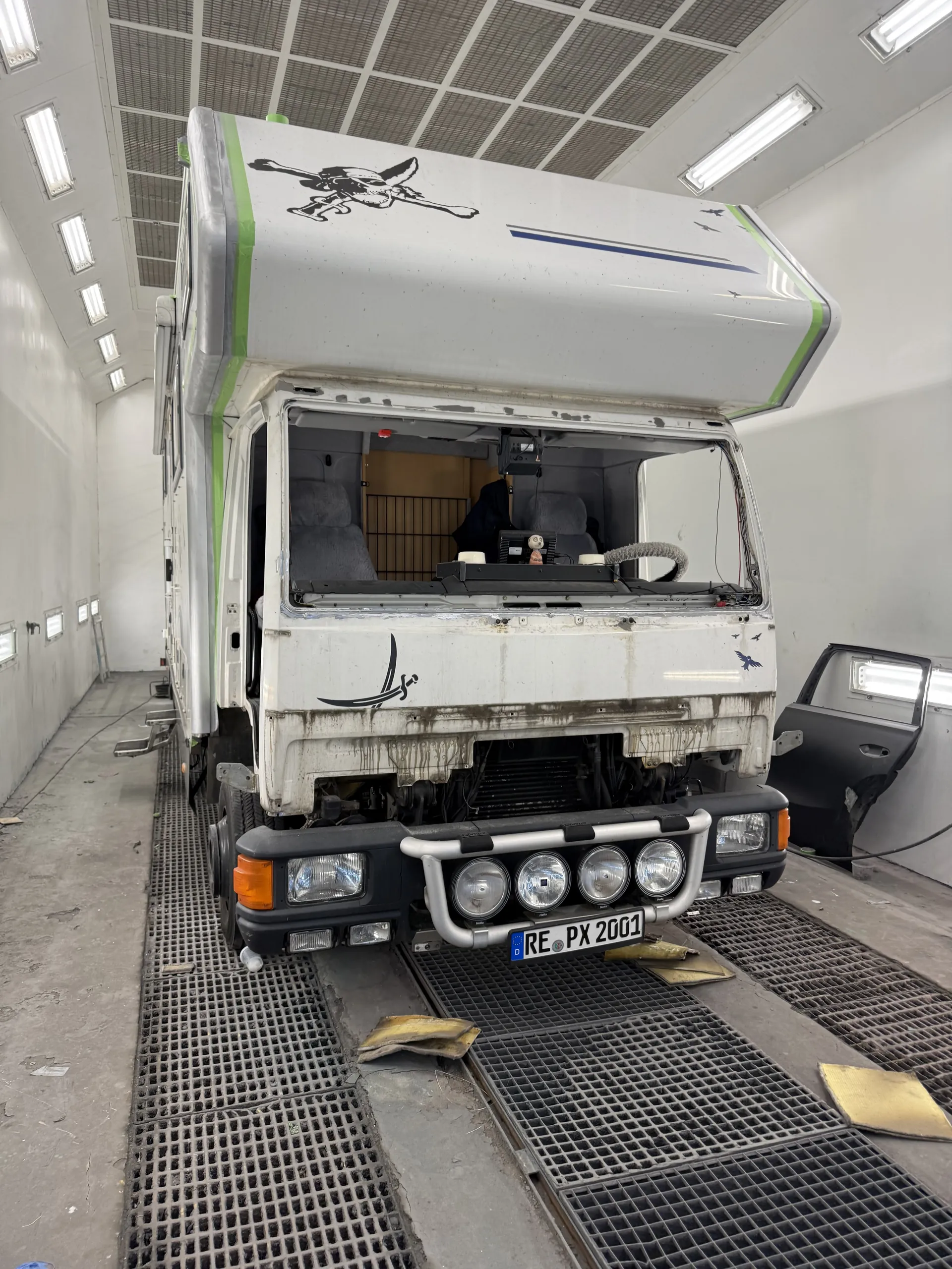 Ein weißes Wohnmobil mit Totenkopf-Logo steht ohne Frontscheibe in einer Lackierkabine.