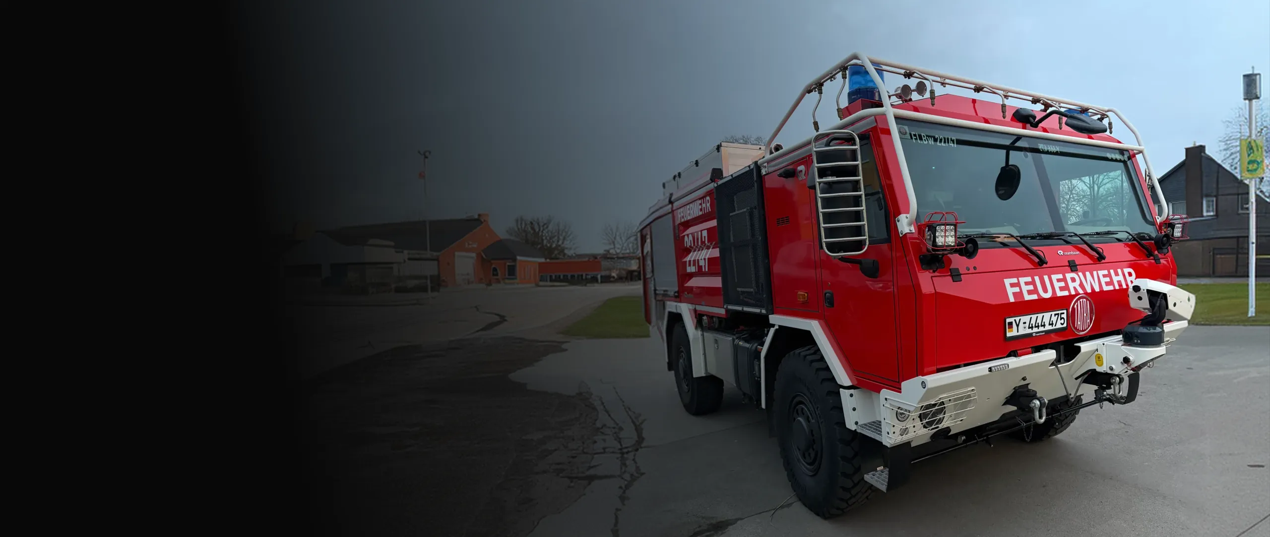 Rotes Feuerwehrfahrzeug mit weißer Aufschrift FEUERWEHR auf einem asphaltierten Platz vor bewölktem Himmel.
