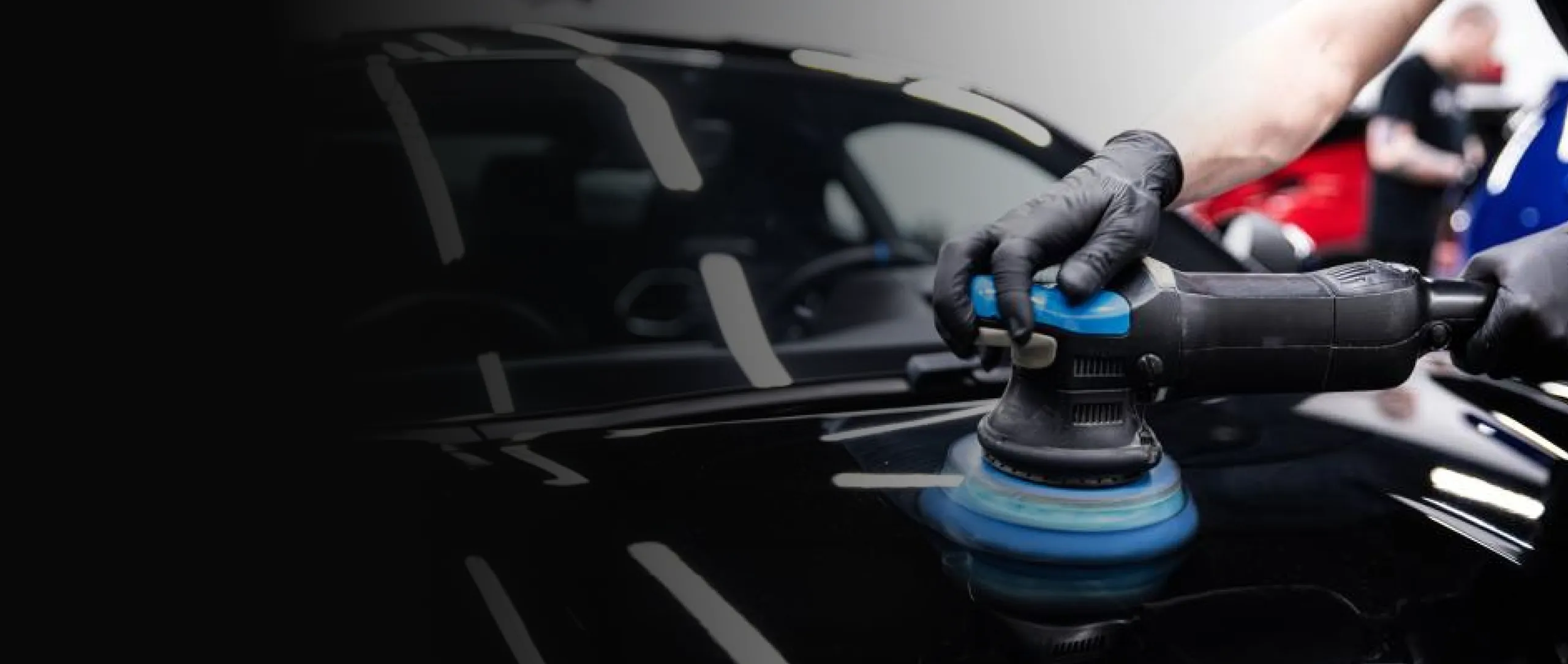 Hände in schwarzen Handschuhen führen eine Poliermaschine über den glänzenden schwarzen Lack eines Autos.
