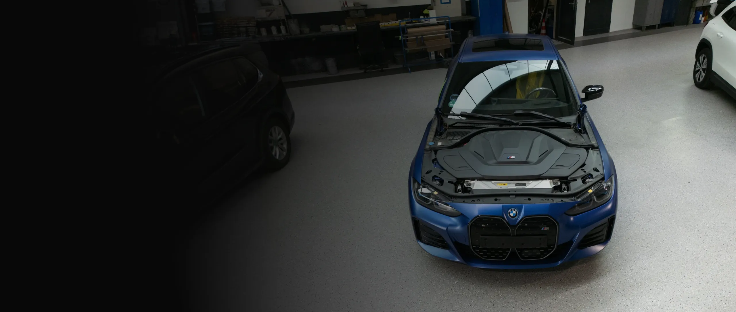 Beschädigter grauer BMW mit Frontschaden steht neben weiteren Fahrzeugen in einer Werkstatt oder Garage.